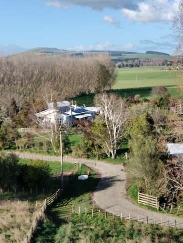 Cette image montre une ferme rurale entourée d'arbres et de champs verdoyants, traversée par une allée de gravier. Les collines et le ciel bleu dominent la scène paisible de la campagne.