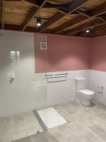 This bathroom features a modern design with light pink walls, tiled flooring, exposed wooden ceiling beams, a glass shower, white toilet, towel rack, and minimalistic fixtures.