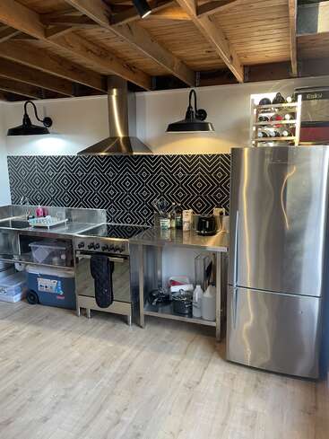 This modern kitchen features stainless steel appliances, geometric black-and-white backsplash, open shelving, exposed wooden ceiling, bright lighting, and a wooden wine rack on the fridge.