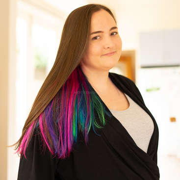The image depicts a young woman with long brown hair featuring vibrant pink, blue, and green highlights, wearing a black cardigan over a light-colored shirt.
