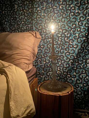 Une bougie allumée sur un support en verre est posée sur une souche en bois à côté d'un lit avec des draps beiges, devant un rideau décoratif à motifs bleus.