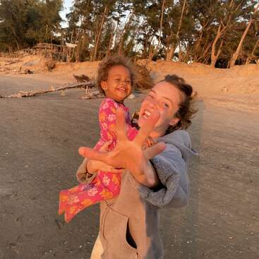 Une femme tient joyeusement un enfant souriant sur une plage de sable, en s'approchant de l'appareil photo. La lumière dorée du soleil, les grands arbres et le bois flotté créent une atmosphère chaleureuse et ludique.