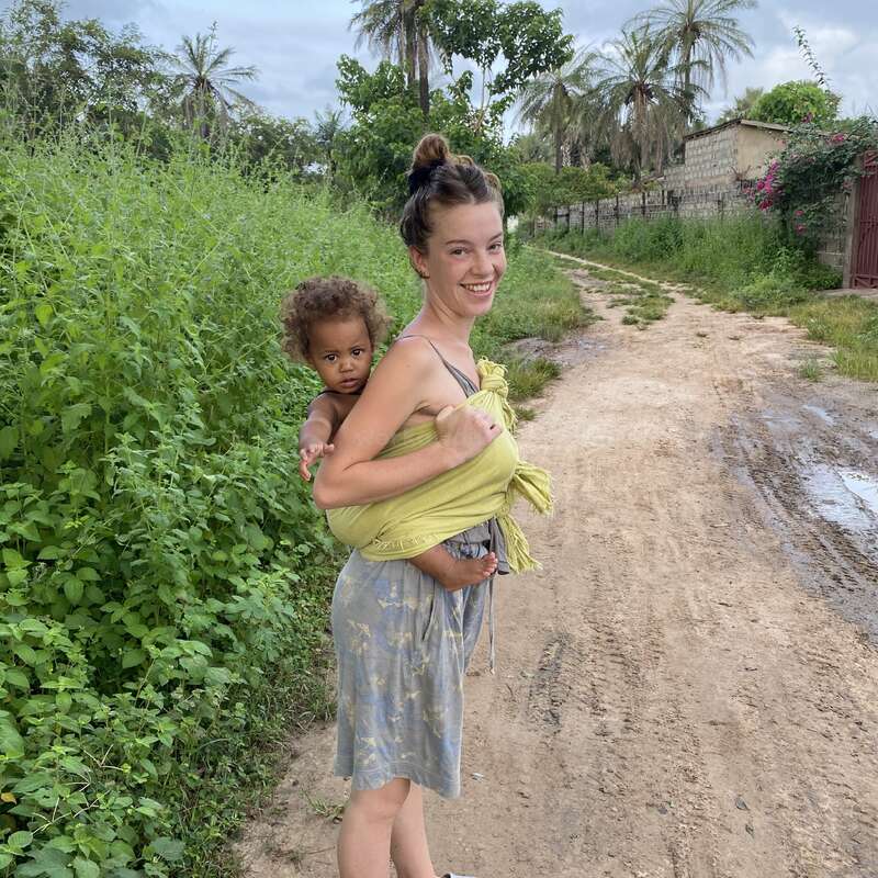 Une femme souriante se tient sur un chemin de terre, portant un jeune enfant sur son dos dans une écharpe verte. Des palmiers et de la verdure luxuriante les entourent.