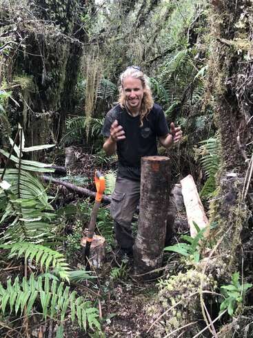 Ein Mann mit langen blonden Haaren steht in einem dichten, grünen Wald. Er lächelt neben gefällten Baumstämmen und einer Säge, umgeben von üppigen Farnen und moosbewachsenen Bäumen.