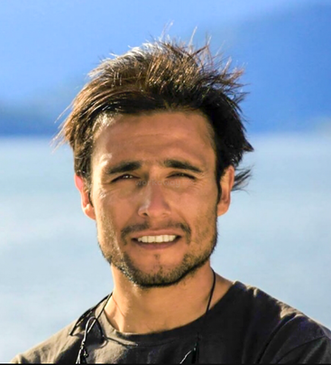 Ein Mann mit zerzaustem dunklem Haar und Bartstoppeln lächelt leicht. Er trägt ein dunkles Hemd und hat eine Sonnenbrille um den Hals gehängt; er steht an einem sonnigen Tag draußen am Wasser.