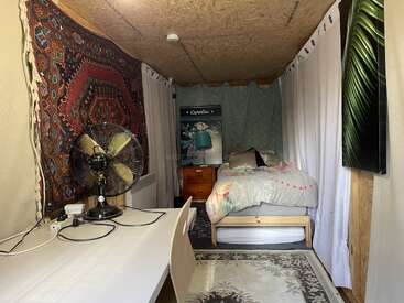 L'image représente une petite pièce avec un lit, un bureau et un ventilateur, avec un mélange de motifs et de textures, y compris une tapisserie, un tapis et des rideaux.
