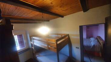 This cozy bedroom features wooden bunk beds, a slanted wooden ceiling, warm lighting, a small window, and an adjacent room with a larger bed visible through the doorway.