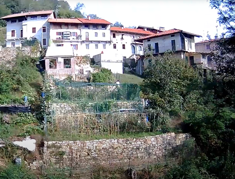 A cluster of white buildings with red roofs sits on a hillside. Terraced gardens and greenery surround the homes, with stone retaining walls and sunlight bathing everything.