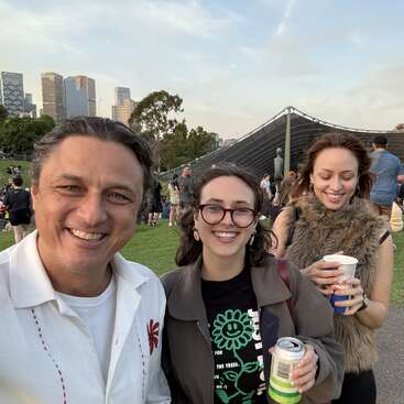 The image depicts three individuals posing for a selfie in an outdoor setting, with a city skyline and grassy area in the background, featuring a large building or structure.