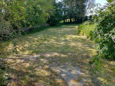 A sunlit grassy path bordered by trees and bushes, with dappled shade covering parts of the ground. The area looks peaceful and secluded, perfect for relaxation.