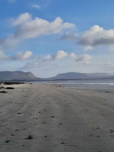 A wide, empty sandy beach stretches into the distance. Gentle waves meet the shore. Mountains rise in the background, under a bright sky with scattered clouds.