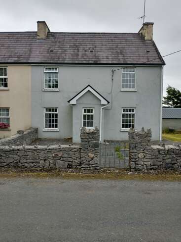 A quaint grey house with white trim, slate roof, and two chimneys, is enclosed by a rustic stone wall and metal gate, under a cloudy sky.