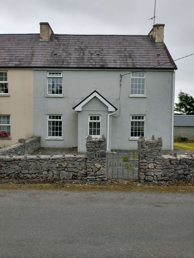 A quaint grey house with white trim, slate roof, and two chimneys, is enclosed by a rustic stone wall and metal gate, under a cloudy sky.