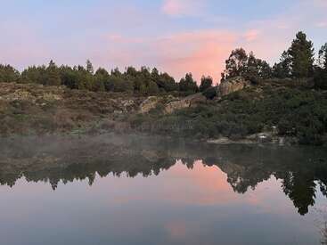Un lago tranquilo refleja colinas rocosas y pinos bajo un cielo rosa y azul al atardecer. La bruma se cierne suavemente sobre el agua, realzando la serenidad.