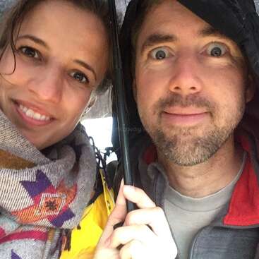 Das Bild zeigt einen Mann und eine Frau unter einem Regenschirm, die in die Kamera lächeln, wobei die Frau auf der linken und der Mann auf der rechten Seite steht.