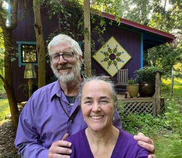 A smiling older couple stands in front of a colorful purple cabin decorated with a large quilt pattern, surrounded by trees and lush, green nature.