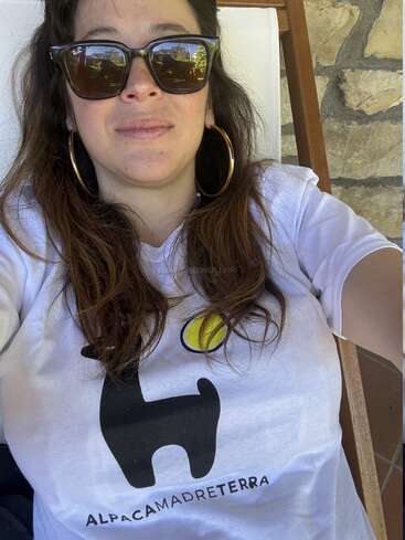 A woman wearing sunglasses and large hoop earrings relaxes outdoors. She wears a white t-shirt with a black alpaca graphic and the words "ALPACAMADRETERRA."