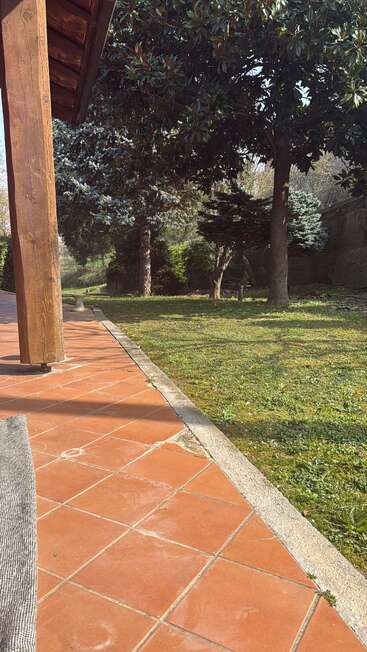 The image shows a sunlit patio with terracotta tiles, bordered by grass and large trees, creating a serene outdoor space under a wooden-roofed structure.