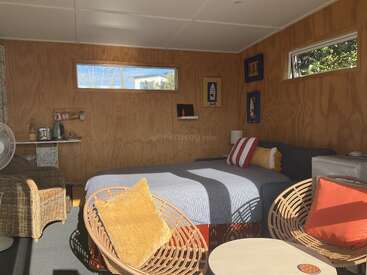 Este quarto aconchegante tem uma cama confortável com almofadas coloridas, paredes de madeira, cadeiras de vime, janelas pequenas, uma mesinha lateral, um abajur e uma cozinha compacta.