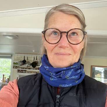 Uma mulher de cabelos grisalhos e óculos está em pé dentro de casa, vestindo um cachecol azul da Nat Geo Adventure, um colete preto e uma blusa laranja. A cozinha fica visível ao fundo.
