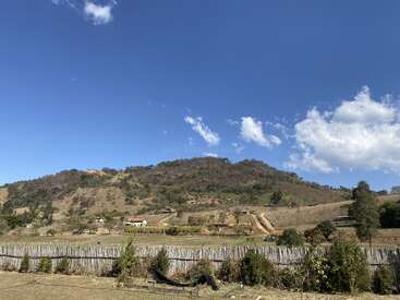 Un paysage pittoresque avec des collines, des arbres clairsemés et des maisons éparses sous un ciel bleu vif avec quelques nuages, clôturé par des piquets de bois et de la verdure devant.