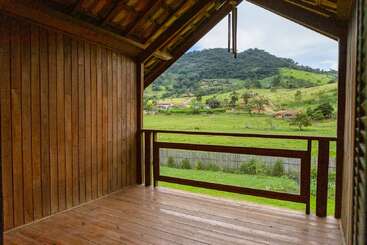 Un balcon en bois avec un toit incliné surplombe un paysage verdoyant avec des collines, des arbres, des maisons éparpillées et une scène rurale paisible sous un ciel nuageux.
