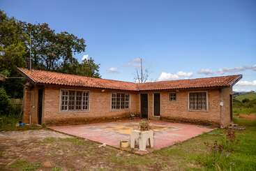 Une maison rustique en briques avec un toit en tuiles rouges se dresse dans une zone rurale, entourée d'herbe et d'arbres, avec une grande cour et des meubles d'extérieur éparpillés.