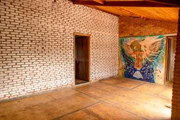 Cette image montre une pièce vide avec des murs en briques blanches, un plafond en bois, une porte et une peinture murale colorée d'une figure mystique sur un mur.