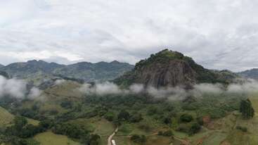 A lush green landscape features a prominent rocky hill, surrounded by mist and scattered trees, with distant mountains and cloudy skies creating a serene atmosphere.