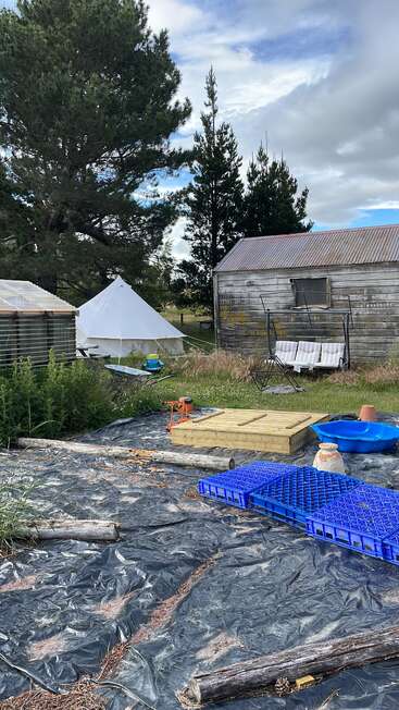 A rustic backyard scene with a weathered shed, tent, garden swing, pallets, raised bed, trees, scattered tools, and tarpaulin covering the ground under cloudy skies.