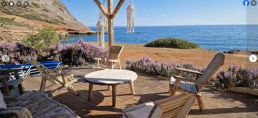 Wooden chairs and table sit on a deck overlooking the sea. Lavender bushes, rocky coastline, and clear blue sky create a tranquil, inviting atmosphere.