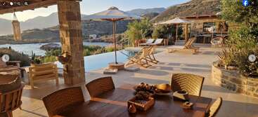 This serene outdoor patio features a pool, sun loungers, umbrellas, rustic furniture, and breathtaking hillside and sea views under golden sunlight, creating a relaxing Mediterranean retreat.