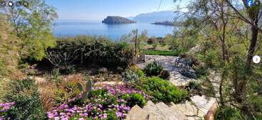 A picturesque seaside garden with vibrant flowers, lush greenery, and stone pathways overlooks calm blue waters and distant islands under a clear, sunny sky. Very serene.