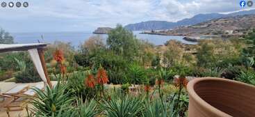 A coastal view with lush green plants and red flowers in the foreground, hills and the sea in the background, under a partly cloudy sky.