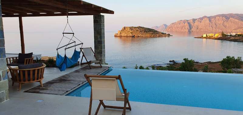 The image depicts a serene poolside scene with a wooden deck, hammocks, and lounge chairs overlooking the ocean, creating a peaceful atmosphere.