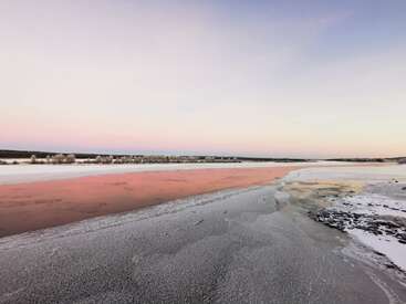A serene winter landscape with a frozen lake, reflecting pink hues from the setting sun. Snow covers the ground, blending gently with the pastel-colored sky. Peaceful.