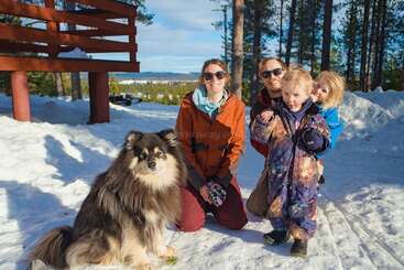 A family with two children and a fluffy dog enjoy a sunny winter day outdoors, surrounded by snow, trees, and blue sky, all smiling happily.