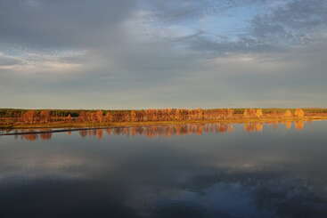 A calm river reflects golden autumn trees and a wide, cloudy sky. The serene landscape evokes tranquility, with distant woods and soft sunlight illuminating the scene.