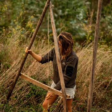 Eine Frau steht im Freien im hohen Gras und hält eine hölzerne Wasserwaage, die für Vermessungen oder Permakultur verwendet wird. Sie wirkt konzentriert, umgeben von üppiger, grüner Natur.