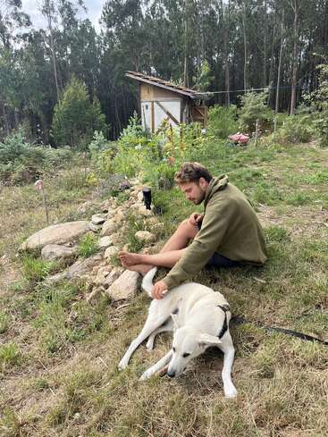 Ein Mann sitzt barfuß im Gras und streichelt einen weißen Hund. Dahinter befindet sich ein rustikaler Schuppen, üppiges Grün und hohe Bäume im Hintergrund. Eine friedliche Szene.