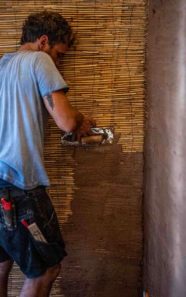 Ein Mann in einem blauen Hemd verputzt eine Wand mit einer Kelle, indem er Lehm über eine Bambuslatte aufträgt und dabei sorgfältig arbeitet.