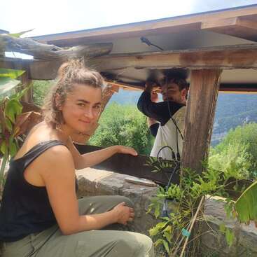 Zwei Menschen arbeiten gemeinsam im Freien an einer Holzkonstruktion. Der eine lächelt in die Kamera, während der andere sich darauf konzentriert, die Kabel oben zu befestigen. Üppiges Grün umgibt sie.