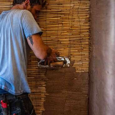 Um homem vestindo uma camisa azul reboca uma parede com uma espátula, aplicando lama sobre uma ripa de bambu, trabalhando cuidadosamente em um ambiente de construção.