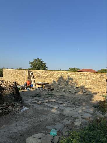 Eine Steinmauer steht unter einem klaren blauen Himmel. Bauwerkzeuge, Baumaterialien, eine Leiter und ein Zementmischer sind zu sehen, dazu verstreute Steine und Erde.