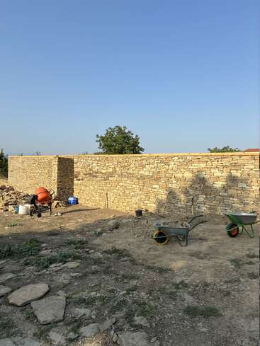 Eine neu errichtete Trockenmauer wird gezeigt, mit Bauwerkzeugen wie Schubkarren und einem Zementmischer im Vordergrund unter einem klaren, blauen Himmel.