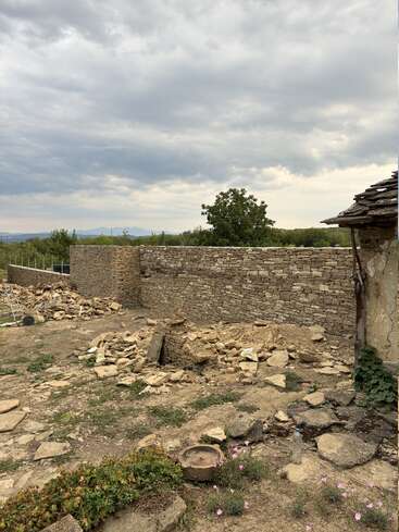 Eine rustikale Steinmauer steht unter einem bewölkten Himmel. Verstreute Steine, Wildblumen und Werkzeuge deuten auf eine laufende Baumaßnahme inmitten einer natürlichen, ländlichen Landschaft mit fernen Bergen hin.