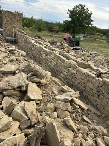 Das Bild zeigt eine teilweise errichtete Steinmauer mit großen Steinen und verstreutem Schutt, Werkzeuge in der Nähe und einen Zementmischer in einem grasbewachsenen Außenbereich.