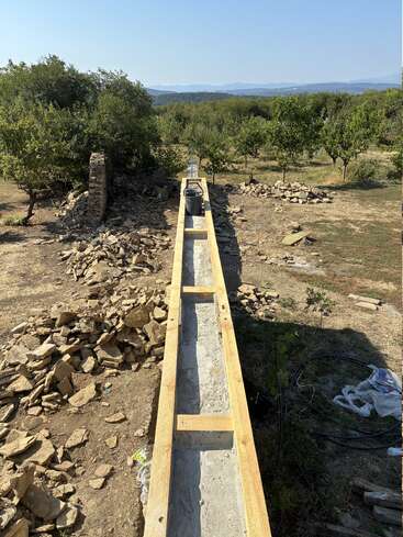 Eine lange Struktur aus Holz und Beton wird im Freien gebaut, umgeben von felsigem Geröll und Bäumen, mit malerischen Hügeln und blauem Himmel im Hintergrund.