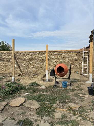 Drei Holzpfähle werden neben einer Steinmauer aufgestellt. Unter einem teilweise bewölkten Himmel sind ein Zementmischer, Werkzeuge, eine Leiter und Gartenpflanzen zu sehen.