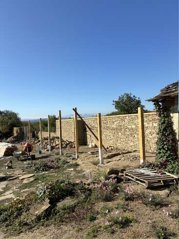 Eine Baustelle mit Holzpfählen, einer Steinmauer, verstreuten Werkzeugen, Blumen und einem klaren blauen Himmel. Einige Kletterpflanzen und eine Holzpalette sind zu sehen.